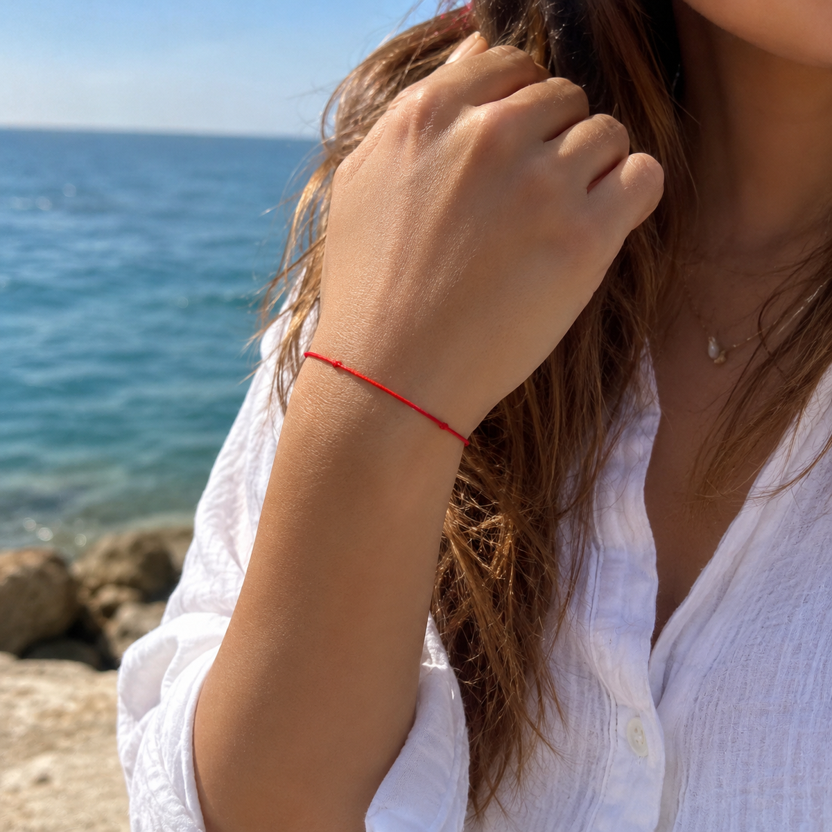 A close-up lifestyle shot of a woman wearing a thin red string bracelet on her wrist, with the ocean and rocks in the background.