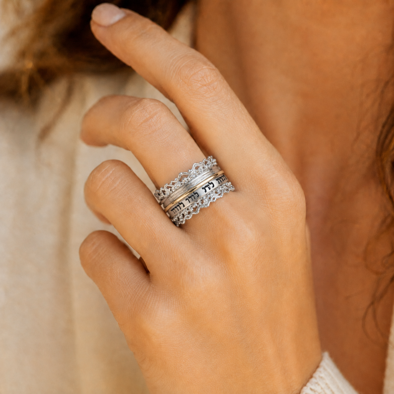 A close-up of a person's hand wearing a silver ring with gold  with intricate filigree and Hebrew lettering, set against a soft beige background.
